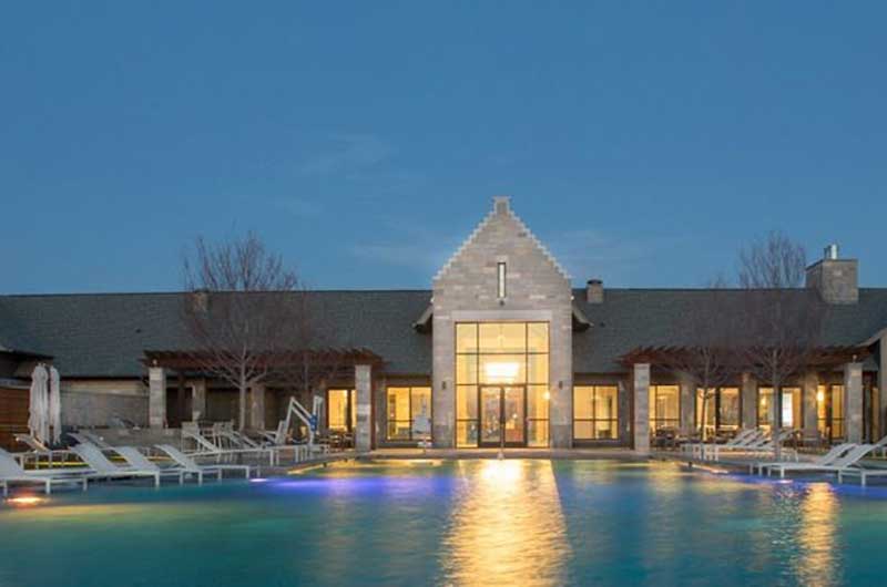 Pool with building behind it at dusk. the building has lights on and the pool is surrounded by lounge chairs.