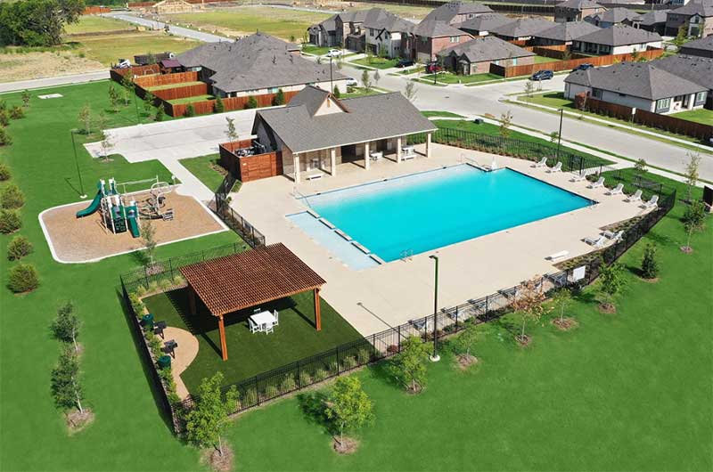 An aerial view of an amenity center with a fenced-in pool and grass and trees surrounding it. There is also a playground and neighborhood in the image outside of the amenity area.