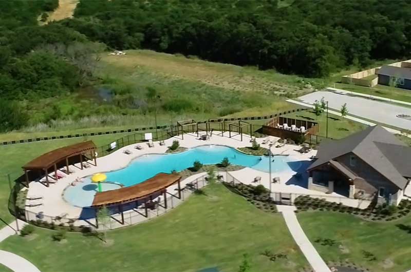 An aerial shot of a pool with pergolas and an amenity center near it. There is also a fence surrounding the pool and a wooded area behind it.